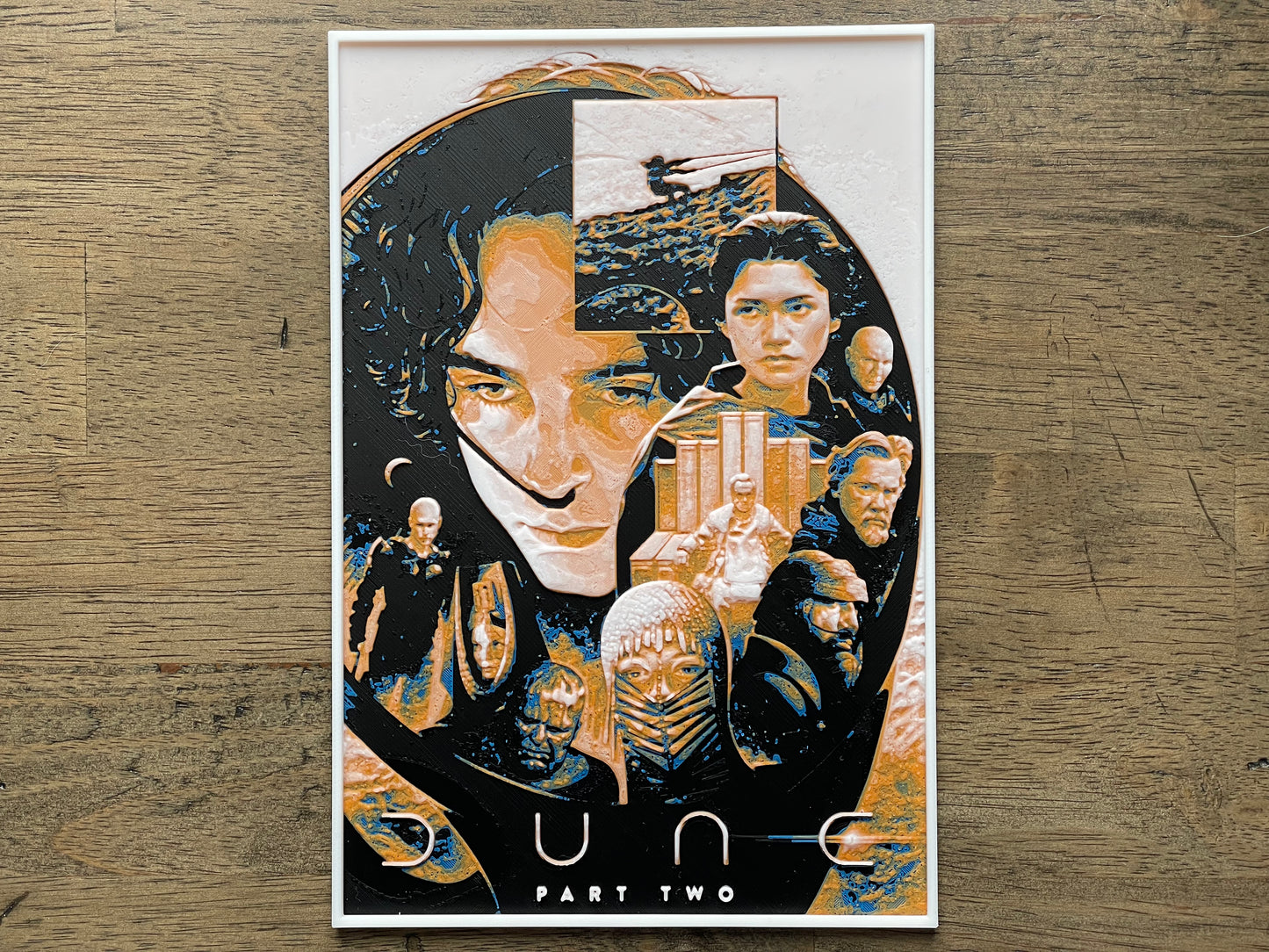 Artistic depiction of a character with multiple faces and a cityscape, titled 'Dune Part Two', on a wooden surface.