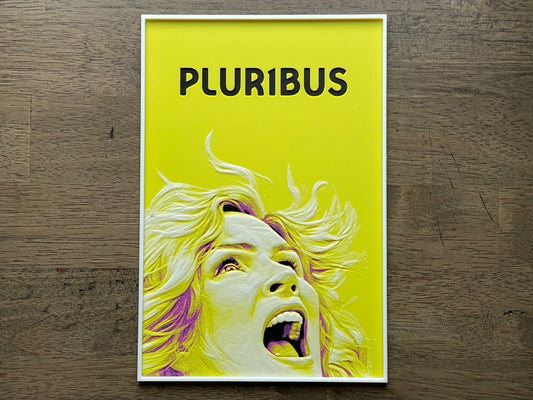 Poster with a stylized face and 'PLURIBUS' text on a yellow background, placed on a wooden surface.
