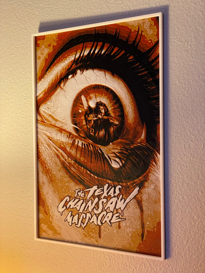 Framed artwork of a stylized eye with 'The Texas Chainsaw Massacre' text on a textured wall.